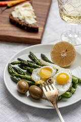 Vertical frame with food: scrambled eggs from two eggs, asparagus, olives, lemon. On a wooden board toast with soft cheese and hot peppers. Next to it is a glass of white wine.