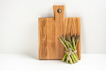 On a white background, a wooden kitchen board and a bunch of fresh green asparagus stand against the wall