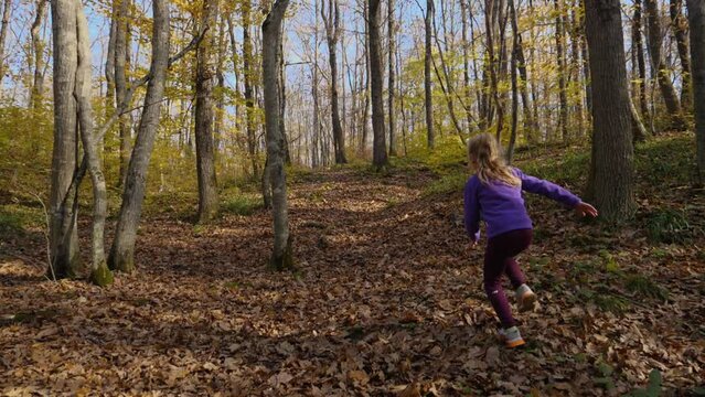 In slow motion, a child girl runs up a hill on dry leaves in the woods. The child runs through the park away from the camera. Active recreation in the woods