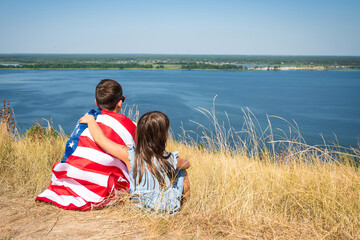 Happy children with the flag of america sit on the bank of the river on a sunny summer day. View from the back. Free lifestyle.