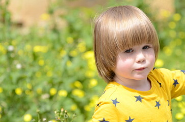 Fototapeta premium Cute positive boy with long blond hair. Close-up portrait against the background of yellow flowers. Place for text