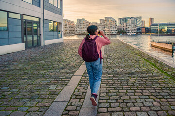 A young girl walks along a pedestrian road with paving stones on the embankment along the water canal and drinks against the backdrop of residential buildings. Copenhagen, Denmark - obrazy, fototapety, plakaty