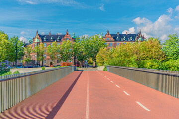Pedestrian and bicycle bridge passes into a park with trees and old brick houses. Copenhagen, Denmark