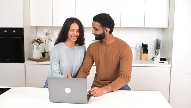 Positive Indian couple in love spends time together at home, using laptop for web surfing, smiling pakistani guy and woman checking emails together, scrolling news, or choosing smth for buying online