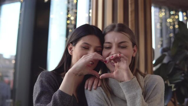 Happy beautiful female gay couple in a cafe shop smiling and making heart shape with their hands,  lesbian love concept