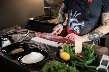 A tattooed chef cooks a juicy steak at home. Beef on a cutting board with a knife and vegetables. Concept of correct cooking of meat by tattoed butcher.