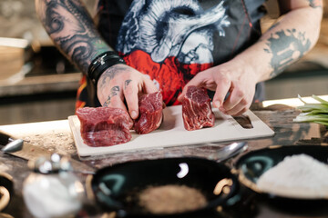 A tattooed chef showing a juicy steak at home. Beef on a cutting board with a knife and vegetables