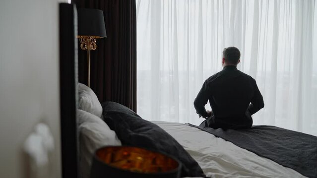 An adult man in a shirt in a room looks out the window. Businessman morning. A cozy room with a bed on which the guy sits with his back. A guy in a black shirt is sitting on the bed.