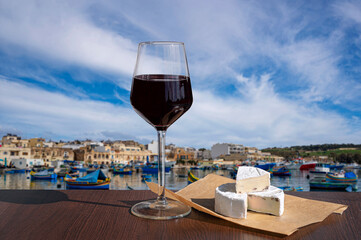 Glass of red wine with brie cheese with view of harbor with traditional boats and historic city center in Marsaxlokk, Malta - obrazy, fototapety, plakaty