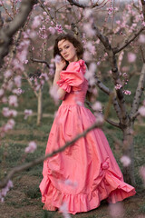 Flowering trees in spring. A beautiful young European girl in a pink dress stands in gardens with peach trees.