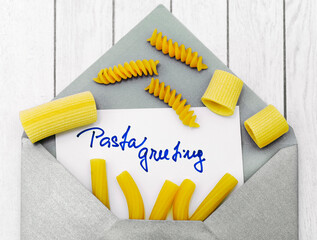 envelope with text pasta greetings and various kinds of raw uncooked pasta over white wood...