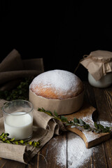 Composition of Easter cake with powdered sugar, a glass of milk, a sieve with powdered sugar in the foreground and a jar of milk in the background on a dark wooden background
