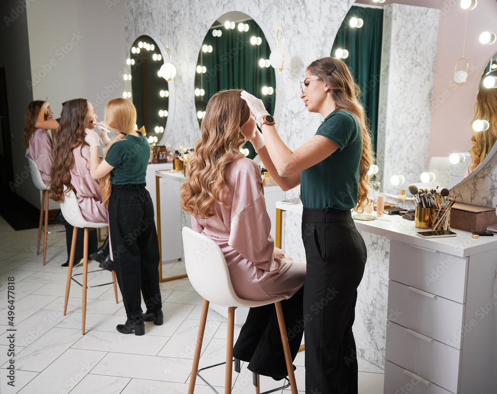 Wall mural Beauty specialists doing professional makeup for clients in modern salon. Young woman with long curly hair sitting at dressing table while female stylist applying foundation with sponge.