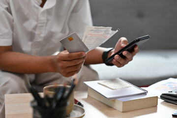 Close up with man sitting on a couch while holding bills and credit card and using mobile phone for calculate, finance, business, home and technology concept.