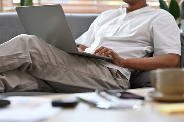 Cropped view man sitting on a couch in a living room looking at a laptop and reading something, for business, education, home and technology concept.