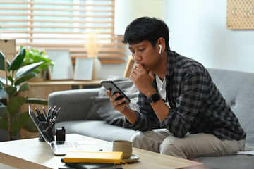 A portrait of a happy Asian man sitting on a couch in a living room using a smart phone and a laptop, for business, communication, home and technology concept.