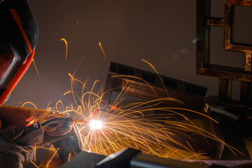A male welder works at home with a semi-automatic welding machine, with golden sparks of steel workpieces. Wear a protective mask and wear protective gloves.