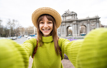Beautiful female tourist taking selfie at Puerta de Alcala in Madrid, Spain - Happy young woman having fun on european city street - Girl smiling at camera outside - obrazy, fototapety, plakaty
