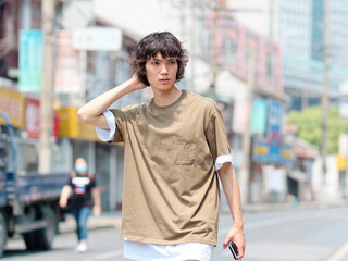 Portrait of handsome Chinese young man with black curly hair in brown T-shirt and pants walking on Shanghai old town street with hand on his head looks lost, front view of modern young people.