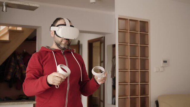 Man At Home Wearing Virtual Reality Headset Holding Gaming Controllers. VR Tester in White Headset Moves Hands With Controllers Man Playing Games On Virtual Reality Device. Metaverse Oculus Technology