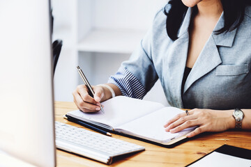 learning, note taking, planning, female employee holding a pen to write a notebook and using a computer to learn how to plan, invest and manage financial risks.