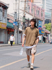 Portrait of handsome Chinese young man with curly black hair in brown T-shirt and pants walking on Shanghai old town street with bottle of water in hand, front view of cool young man.