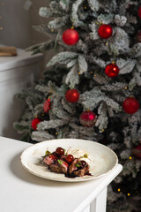 Pieces of stewed beef with cherries in a plate against the background of a decorated Christmas tree