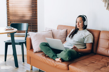 Obraz premium Modern professional woman working in headphones on a laptop while sitting on the couch at home.