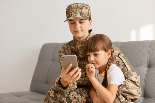 Indoor shot of smiling cam soldier woman wearing camouflage uniform and cap posing with her daughter, looking at cell phone screen, reading news or watching mom's photos from army.
