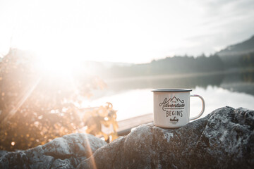 Outdoor mug in front of a lake and forest with blue skies and sun flare in background. The adventure begins cup with travel, hiking and camping concept with copy space