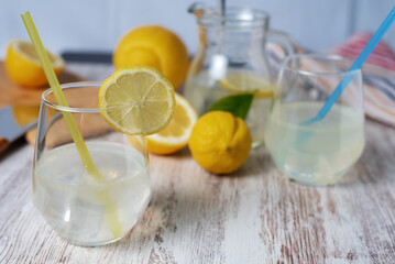 Glass with lemonade and ice, with a slice of lemon on its edge, in the background a pitcher, a cutting board and some out of focus lemons.