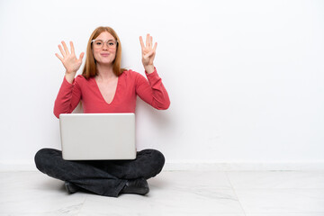 Young redhead woman with a laptop sitting on the floor isolated on white background counting nine...
