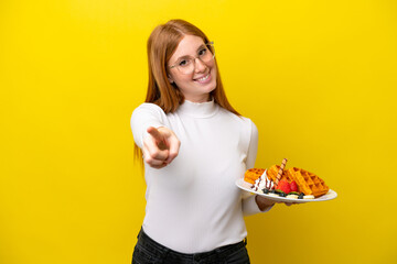 Young redhead woman holding waffles isolated on yellow background pointing front with happy expression