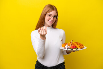Young redhead woman holding waffles isolated on yellow background inviting to come with hand. Happy that you came