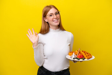 Young redhead woman holding waffles isolated on yellow background saluting with hand with happy expression