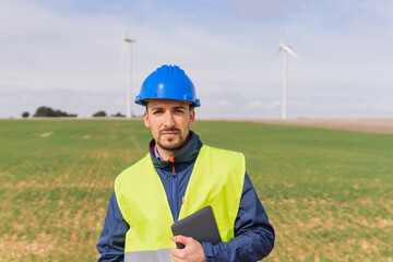 Portrait of a young industrial engineer at a wind turbine farm using his digital tablet. Worker looking at the camera in the facilities of renewable and non-polluting electrical energy.