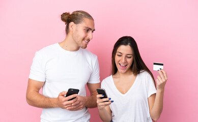 Young caucasian couple isolated on pink background buying with the mobile with a credit card