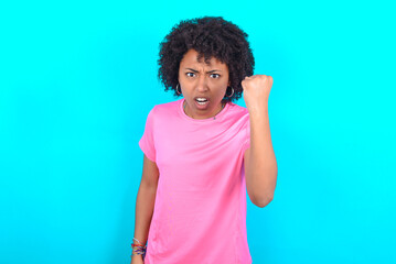 young girl with afro hairstyle wearing pink T-shirt over blue background angry and mad raising fist frustrated and furious while shouting with anger. Rage and aggressive concept.