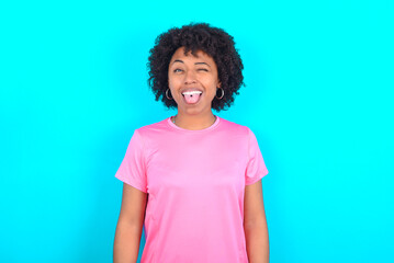 young girl with afro hairstyle wearing pink T-shirt over blue background sticking tongue out happy with funny expression. Emotion concept.