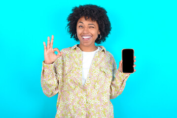 young girl with afro hairstyle wearing floral shirt over blue background holding in hands cell...