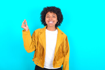 Young woman with afro hairstyle wearing yellow fringe jacket over blue background pointing up with hand showing up seven fingers gesture in Chinese sign language QÄ«.