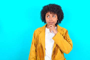 Thoughtful Young woman with afro hairstyle wearing yellow fringe jacket over blue background holds...