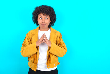 Young woman with afro hairstyle wearing yellow fringe jacket over blue background steepled fingers...