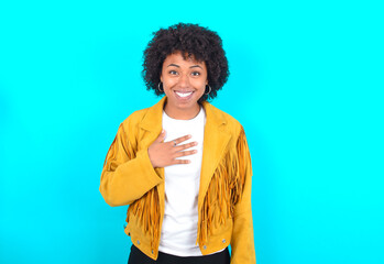 Young woman with afro hairstyle wearing yellow fringe jacket over blue background smiles toothily cannot believe eyes expresses good emotions and surprisement