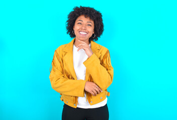 Young woman with afro hairstyle wearing yellow fringe jacket over blue background laughs happily keeps hand on chin expresses positive emotions smiles broadly has carefree expression