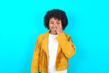 Young woman with afro hairstyle wearing yellow fringe jacket over blue background makes face palm and smiles broadly, giggles positively hears funny joke poses