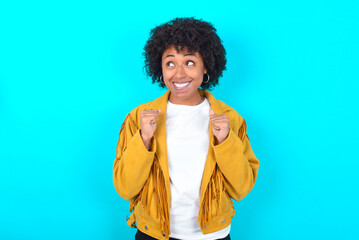 Young woman with afro hairstyle wearing yellow fringe jacket over blue background clenches fists and awaits for something nice happened looks away bites lips and waits announcement of results