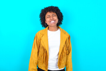 Fototapeta na wymiar Positive Young woman with afro hairstyle wearing yellow fringe jacket over blue background with overjoyed expression closes eyes and laughs shows white perfect teeth