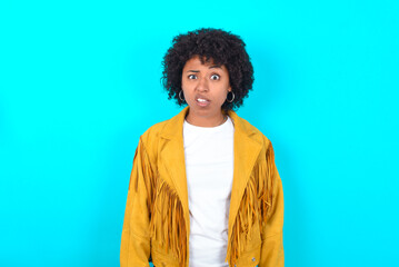 Portrait of dissatisfied Young woman with afro hairstyle wearing yellow fringe jacket over blue...