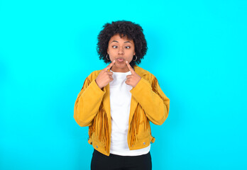 Young woman with afro hairstyle wearing yellow fringe jacket over blue background crosses eyes and...